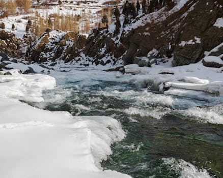 A tranquil stream flowing through a snow-covered valley amidst rocky terrain during winter.