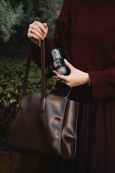 Woman holding a camera and a leather handbag, dressed in a maroon outfit outdoors.