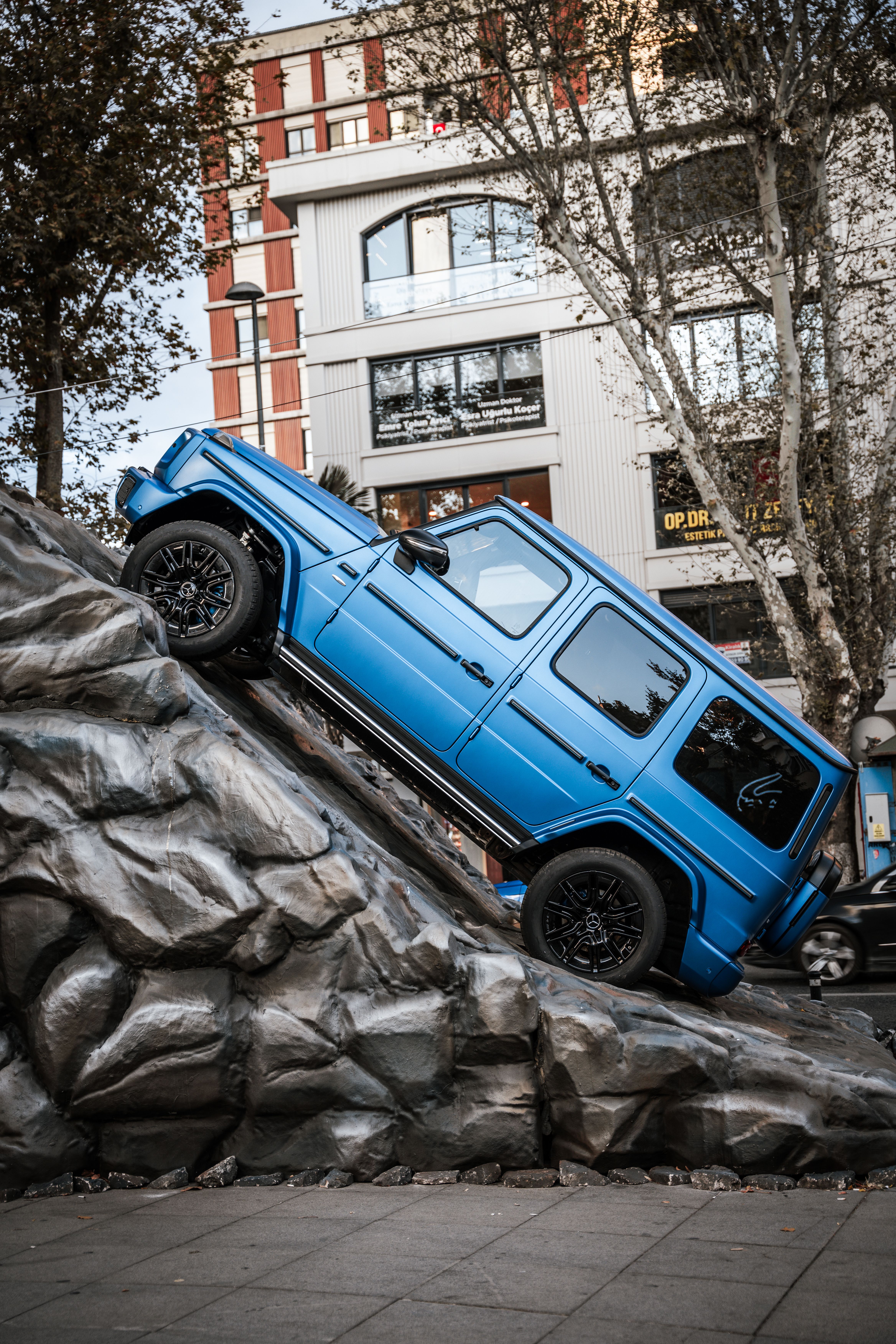 A blue SUV ascends a steep artificial rock in an urban setting in İstanbul, Türkiye.