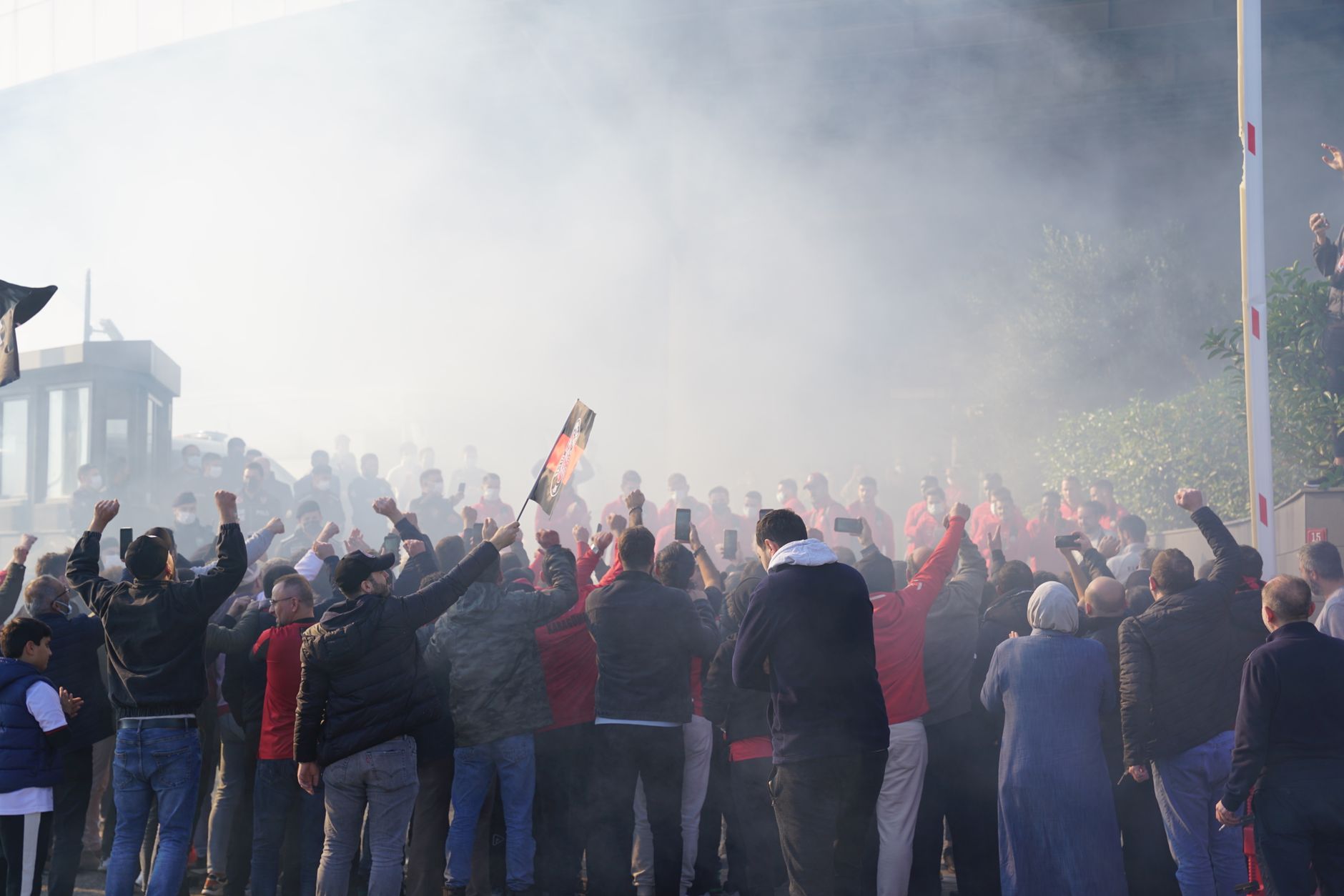 A vibrant group of sports fans showing support with raised arms amidst smoke outdoors.
