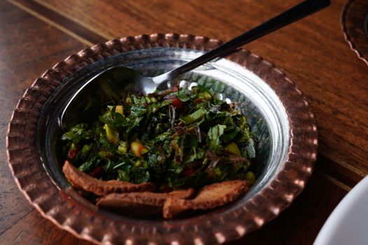 A vibrant tabbouleh salad in a decorative copper bowl with fresh greens and bread on a wooden table.