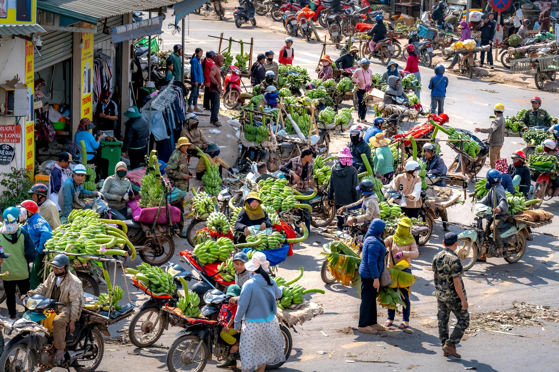 Busy outdoor market with vendors selling bananas on motorcycles.