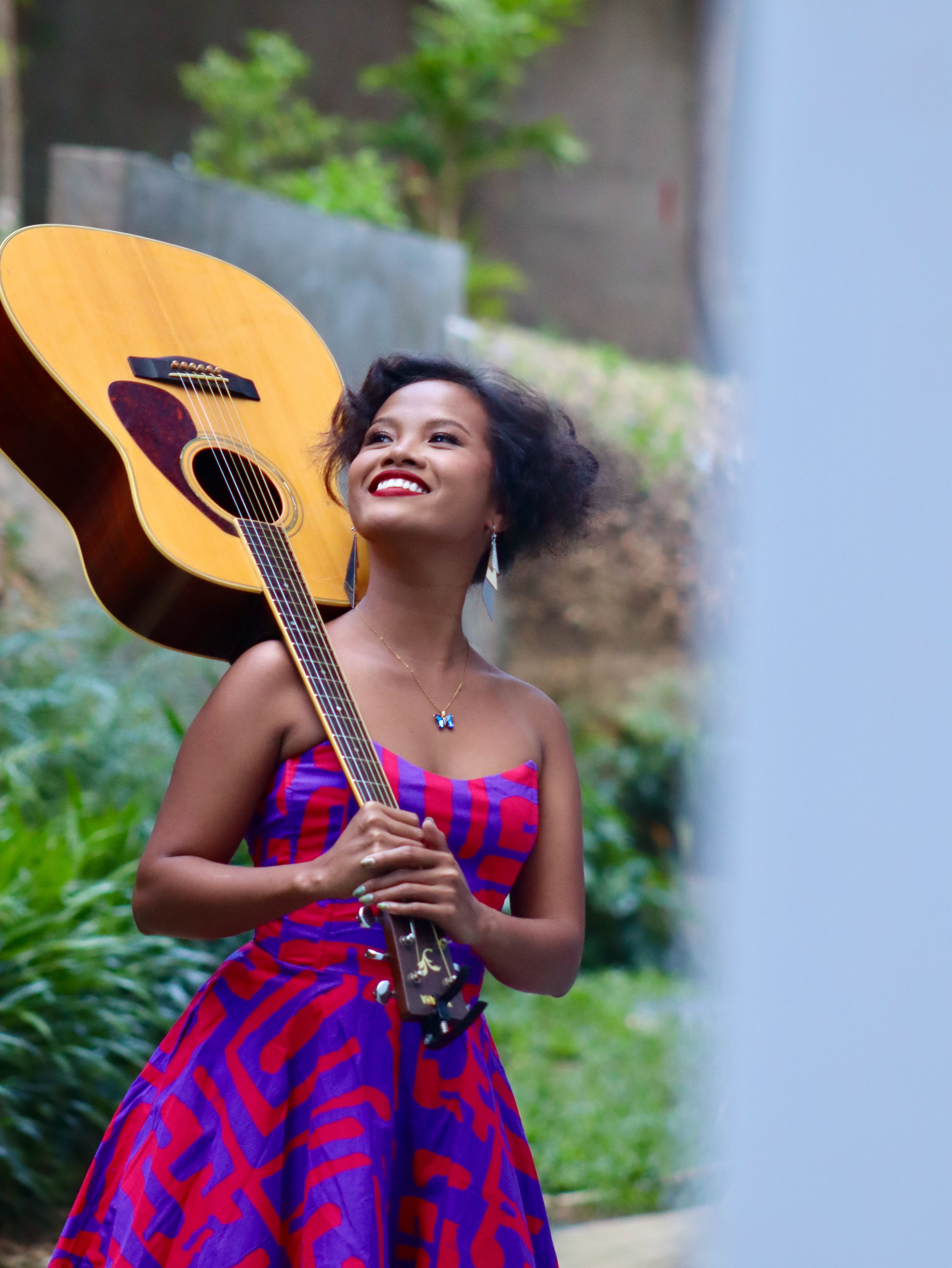 Smiling woman in vibrant dress joyfully holds an acoustic guitar outdoors in Talisay, Philippines.