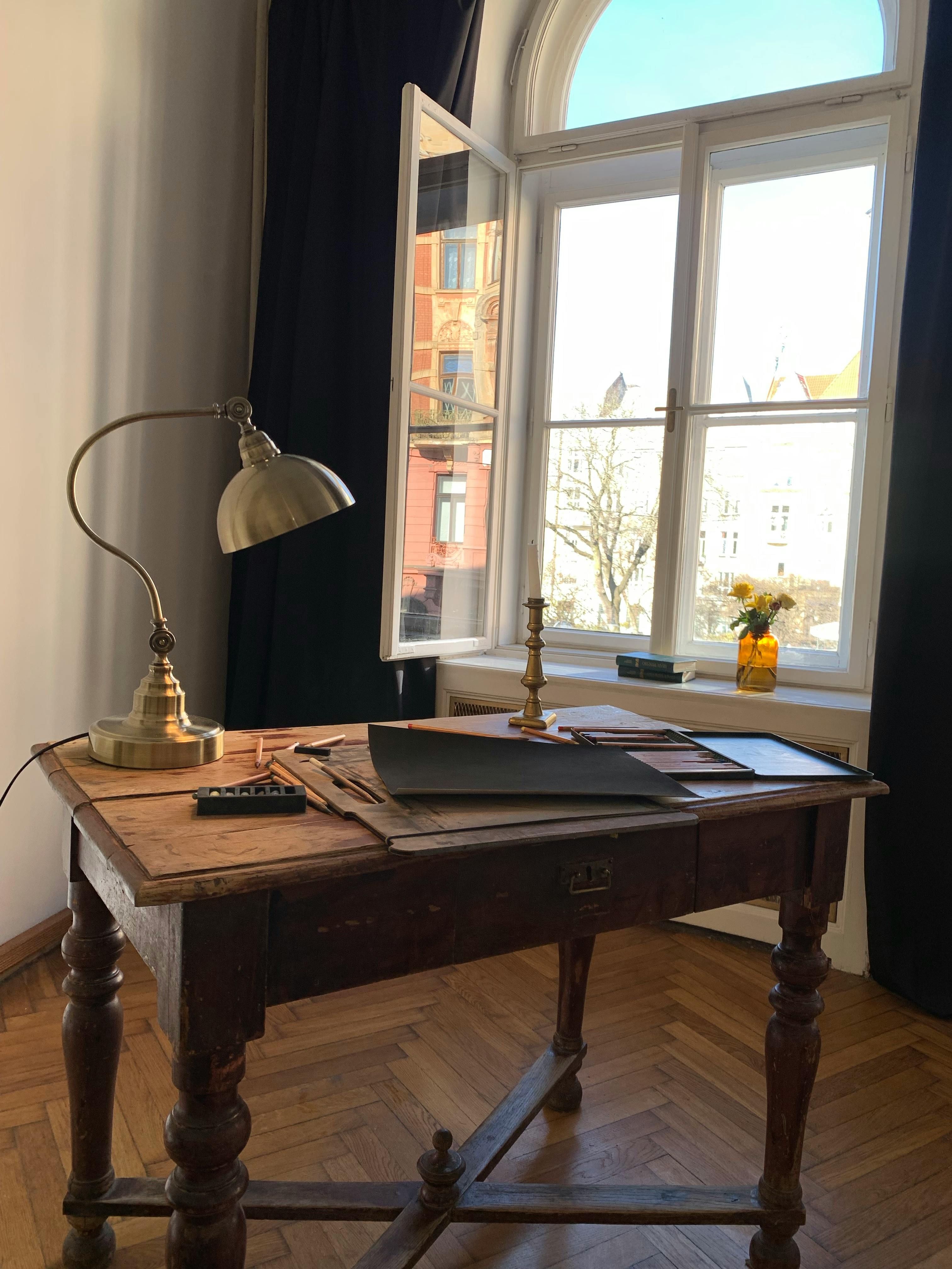 A rustic wooden desk with a lamp and papers by a sunny window.