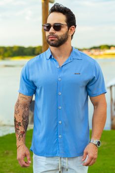 Fashionable man wearing sunglasses and blue shirt outdoors by a lake.