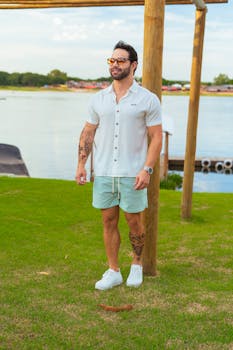 A man wearing a casual summer outfit stands by a tranquil lake, showcasing relaxed style and nature's serenity.