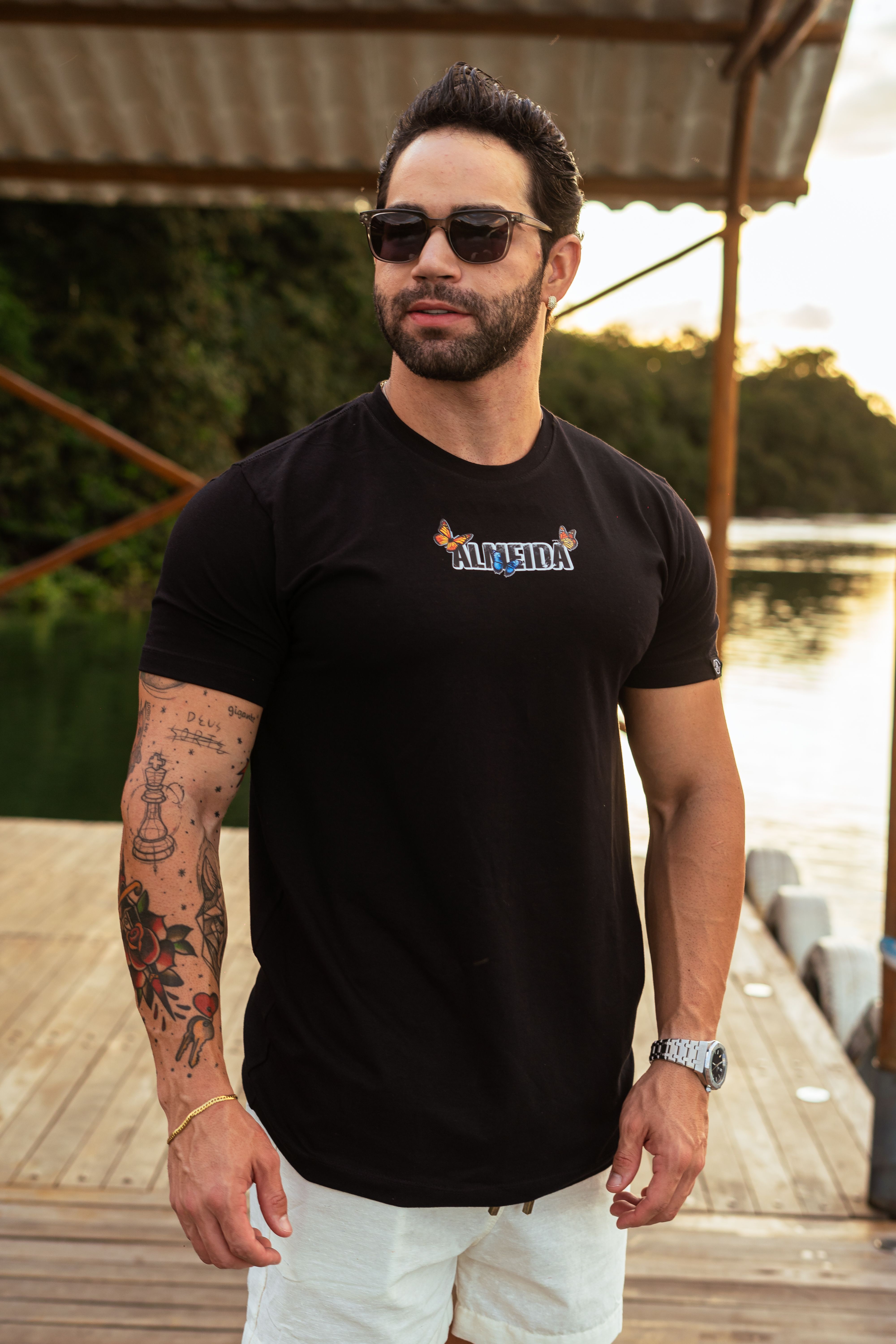 Portrait of a man with tattoos wearing sunglasses on a dock by the water at sunset.