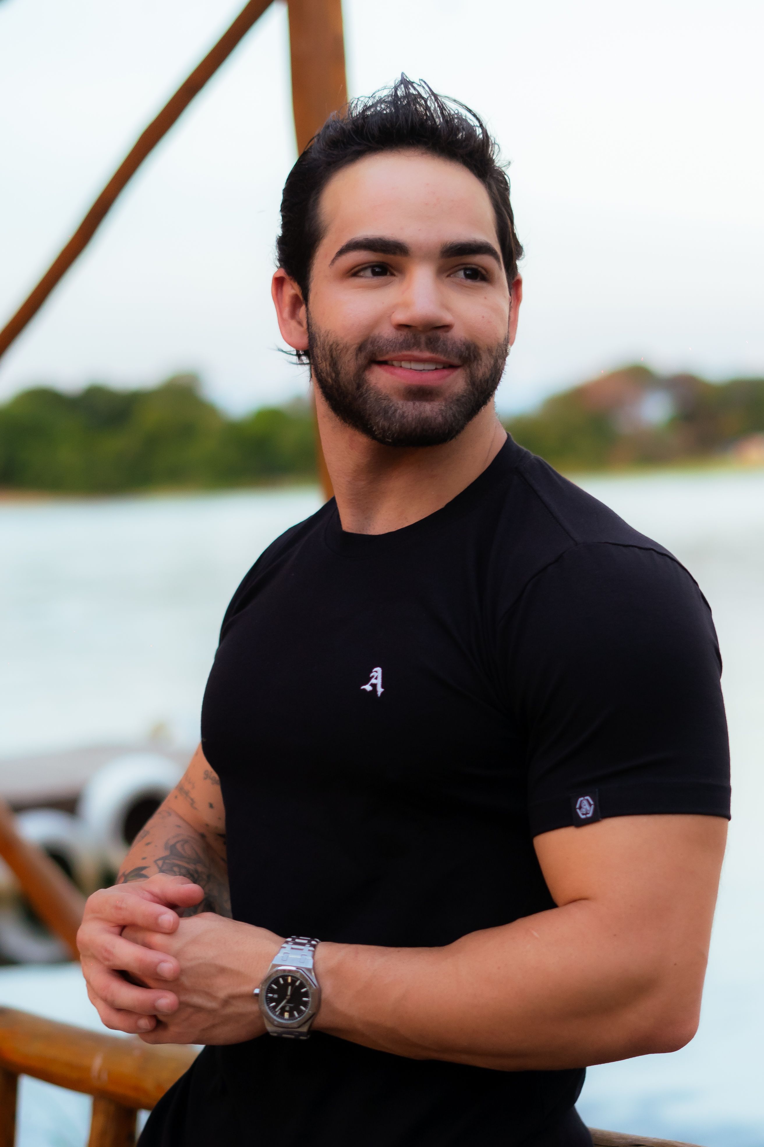 A cheerful man stands by a river, wearing a black shirt and watch, enjoying a daylight moment.