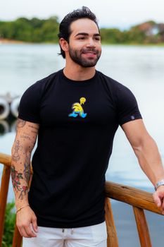 A man with tattoos stands smiling by a scenic riverside in casual attire.