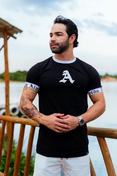 Young man in a black T-shirt with tattoos standing by the lake.
