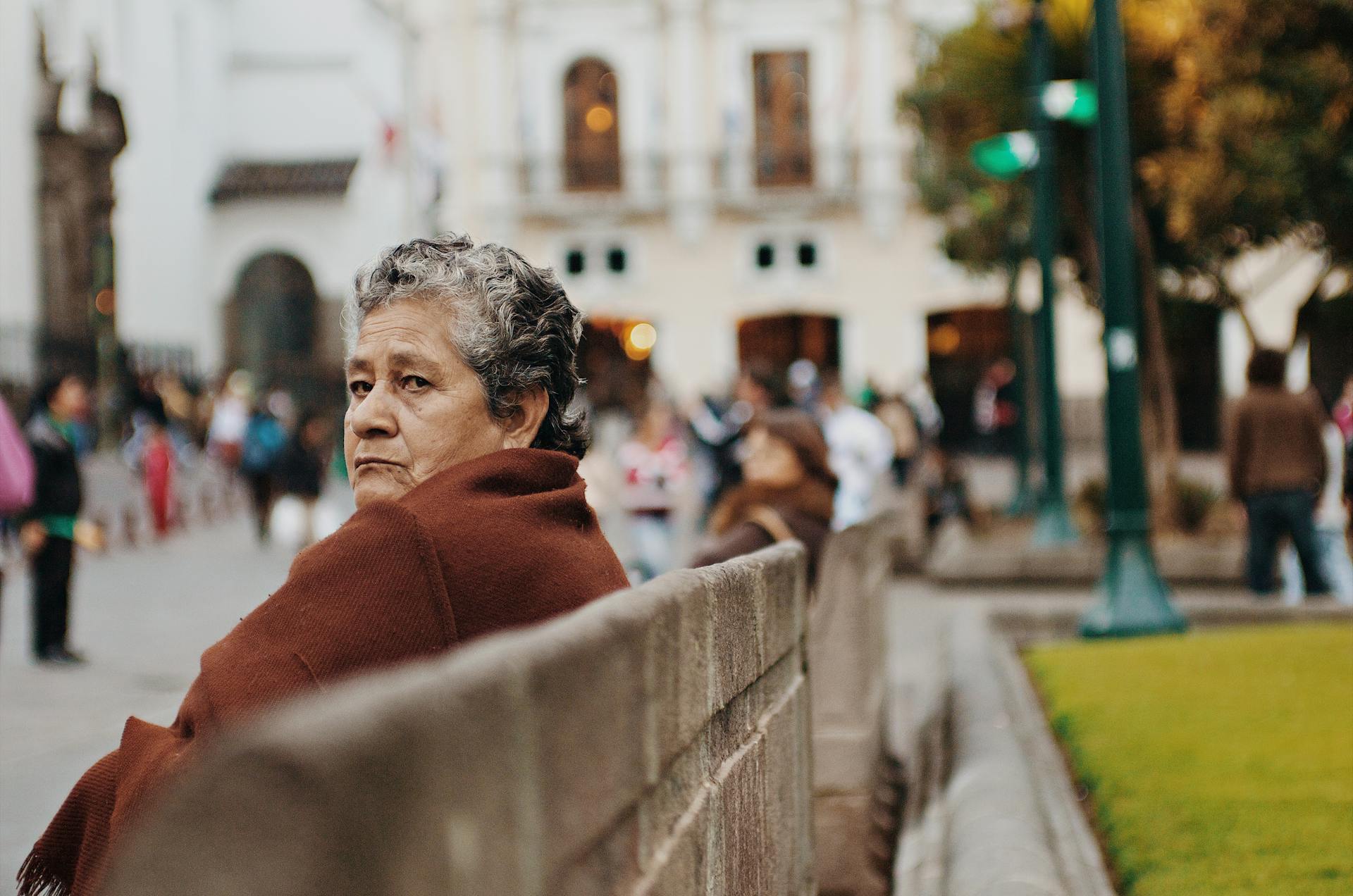 an old, sad, homeless woman sitting on a park bench wearing shawl coat to stay warm