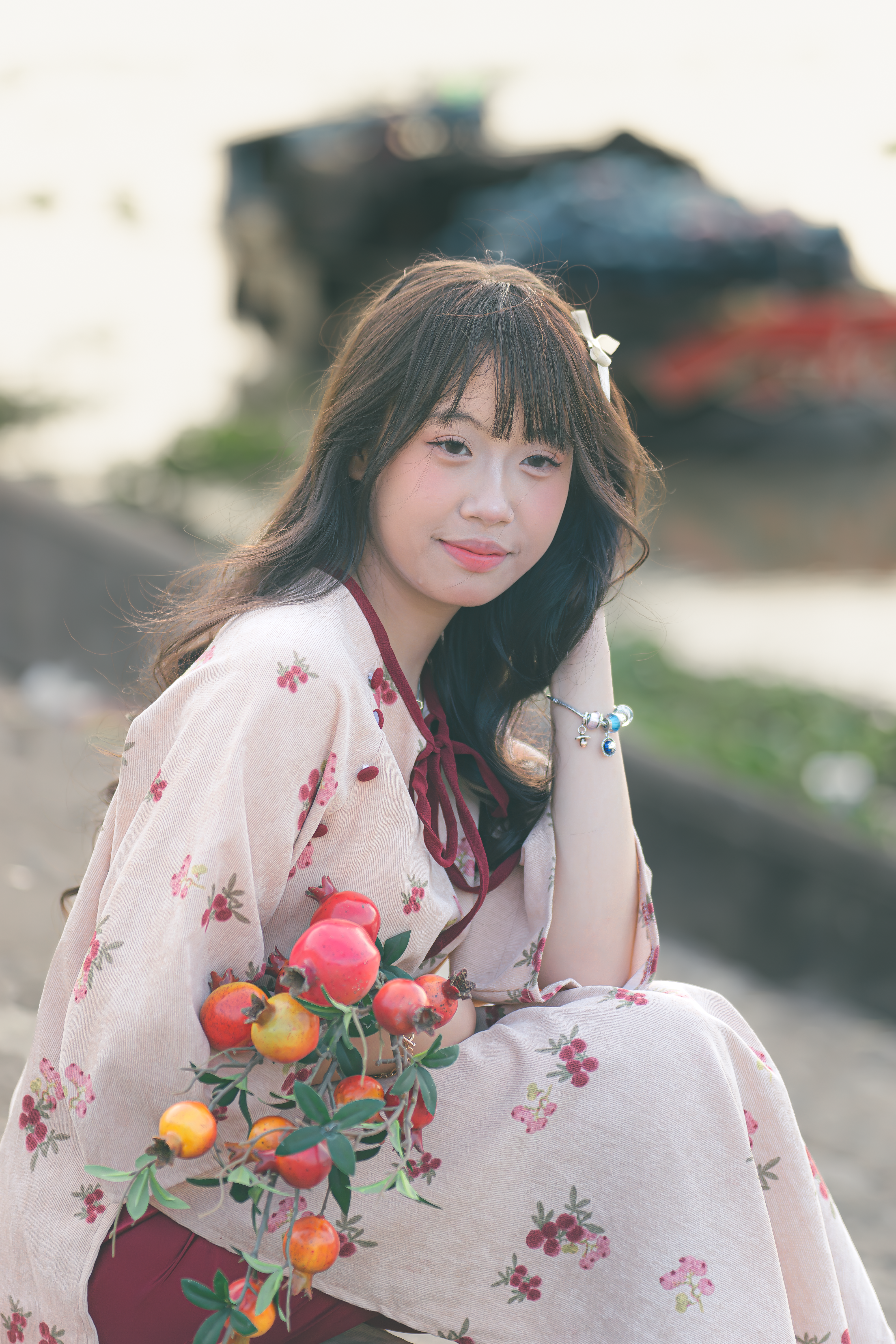 A young woman in a floral dress sits outdoors, holding a fruit bouquet, exuding a peaceful vibe.