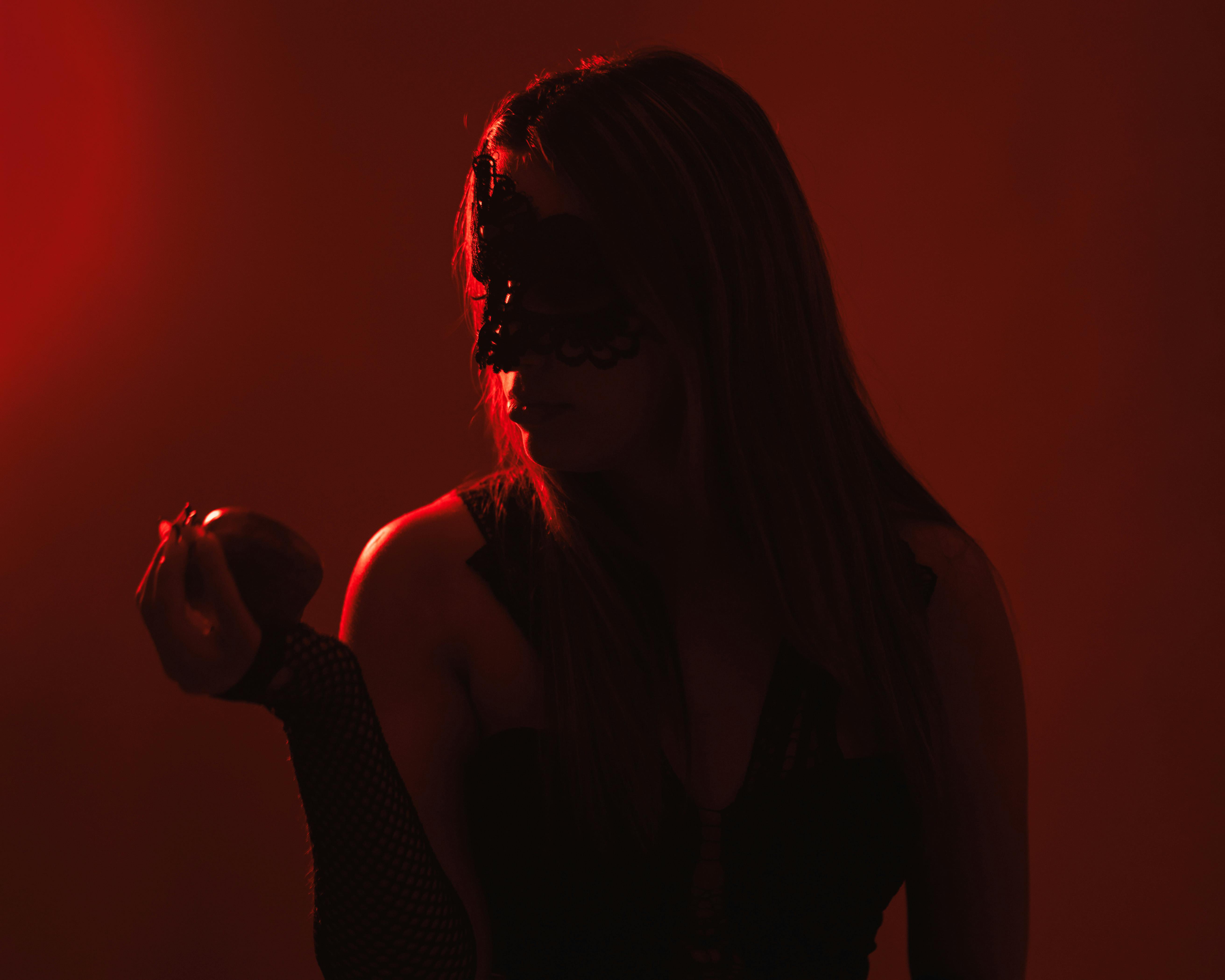 A mysterious woman in a black mask holds an apple under dramatic red lighting.
