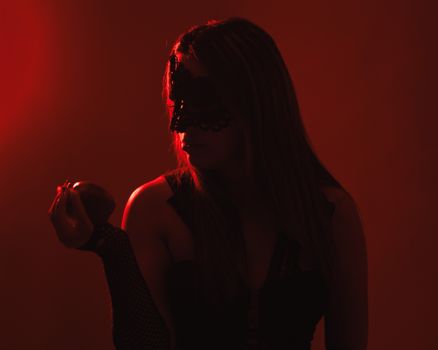 A mysterious woman in a black mask holds an apple under dramatic red lighting.
