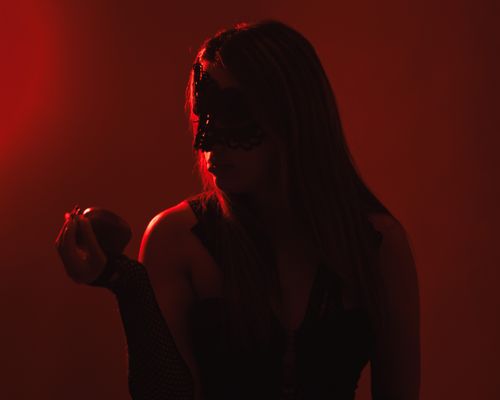 Free A mysterious woman in a black mask holds an apple under dramatic red lighting. Stock Photo