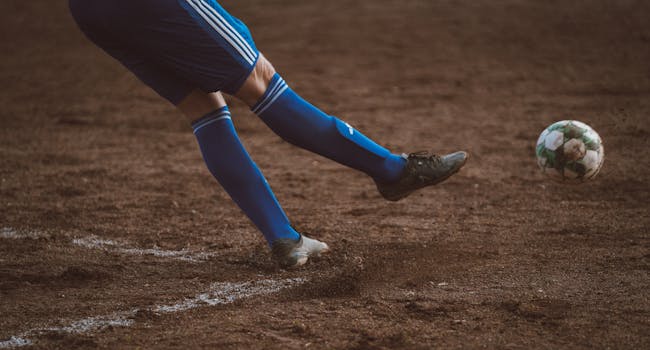 Dynamic soccer player kicks ball on outdoor dirt field. Focus on leg action.