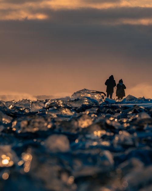Free Two travelers walk at sunset across icy terrain in Iceland, with icebergs glistening in the foreground. Stock Photo