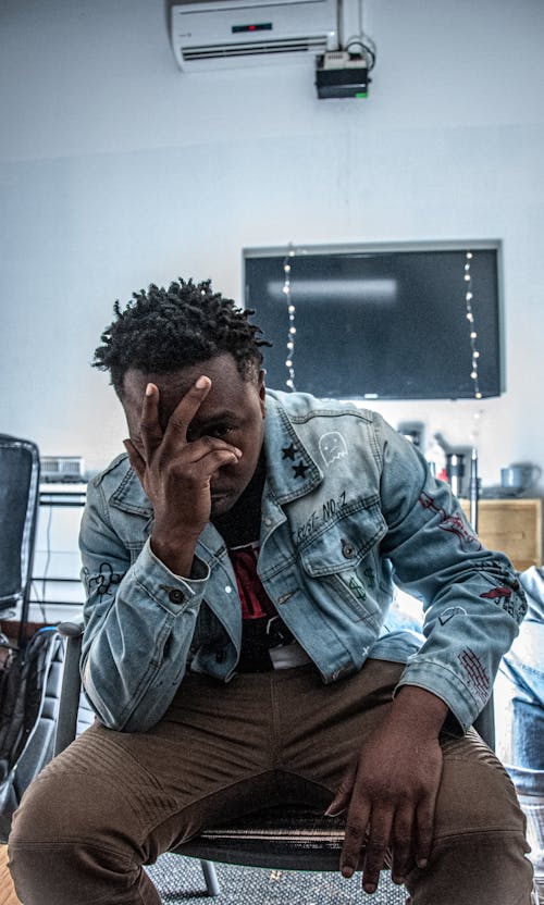 Photo of Sitting Man in Blue Denim Jacket and Brown Pants Posing With His Hands Covering His Face