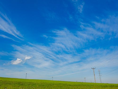 Free Expansive blue sky with light clouds above a wide green field and distant power lines. Stock Photo
