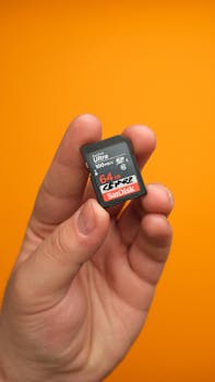 Focused shot of a hand holding a 64GB SanDisk SD card against a bright orange background.