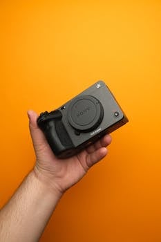 Hand holding a mirrorless camera with an orange backdrop, showcasing sleek design and advanced technology.