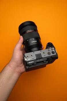 A Sony FX3 cinema camera with a Samyang lens in hand against a bright orange backdrop.
