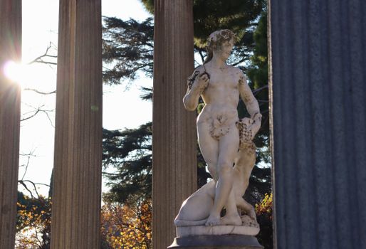 Stone statue of a classical figure amidst columns in a sunny garden.