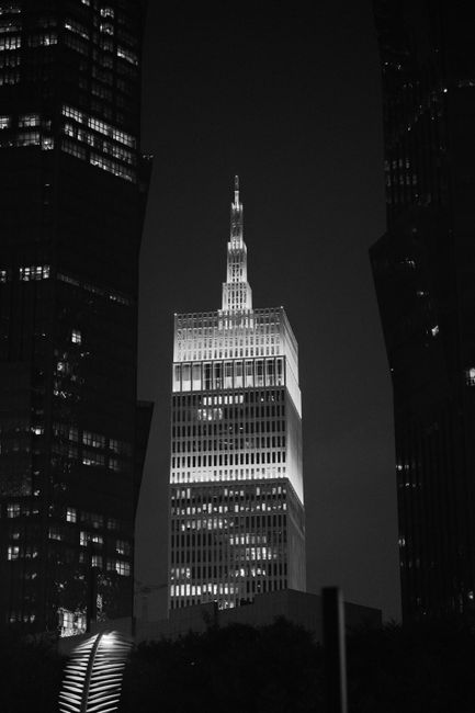 Black and white image of a brightly lit skyscraper against the night sky, framed by taller dark buildings.