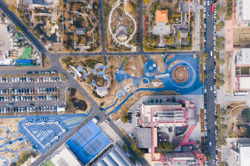 Aerial view of an urban park with playgrounds, parking, and nearby buildings.