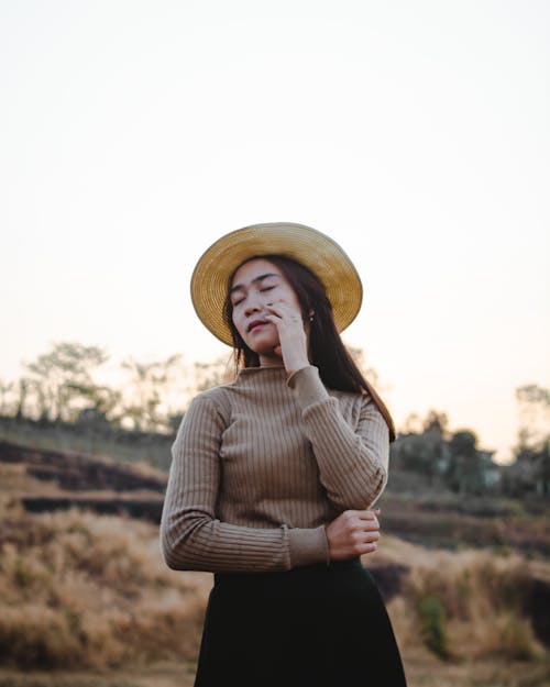 Photo of Woman in Brown Sweater, Black Dress, and Sun Hat Posing With Her Eyes Closed