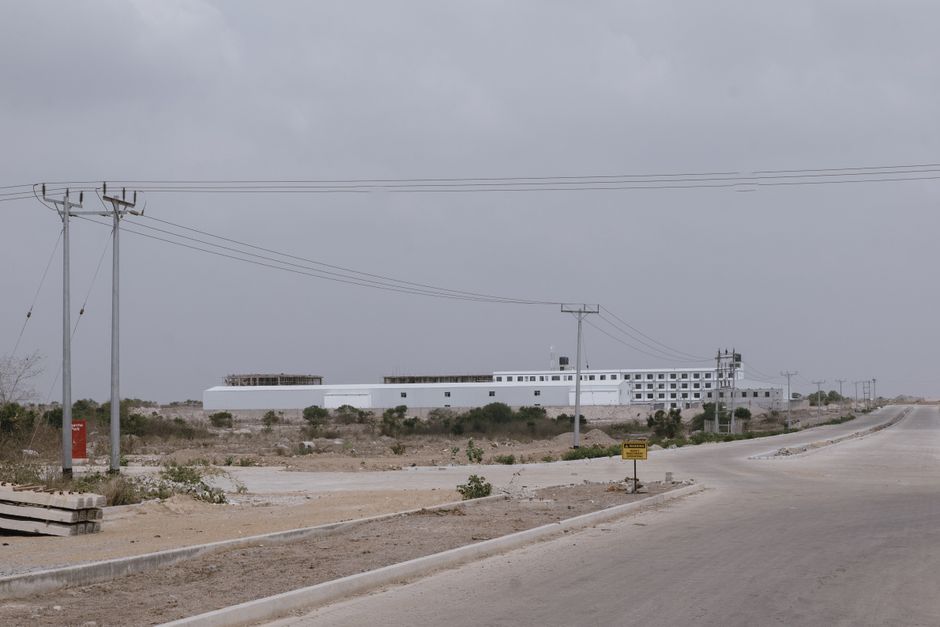 A large industrial building in a barren desert setting under an overcast sky. Power lines run along
