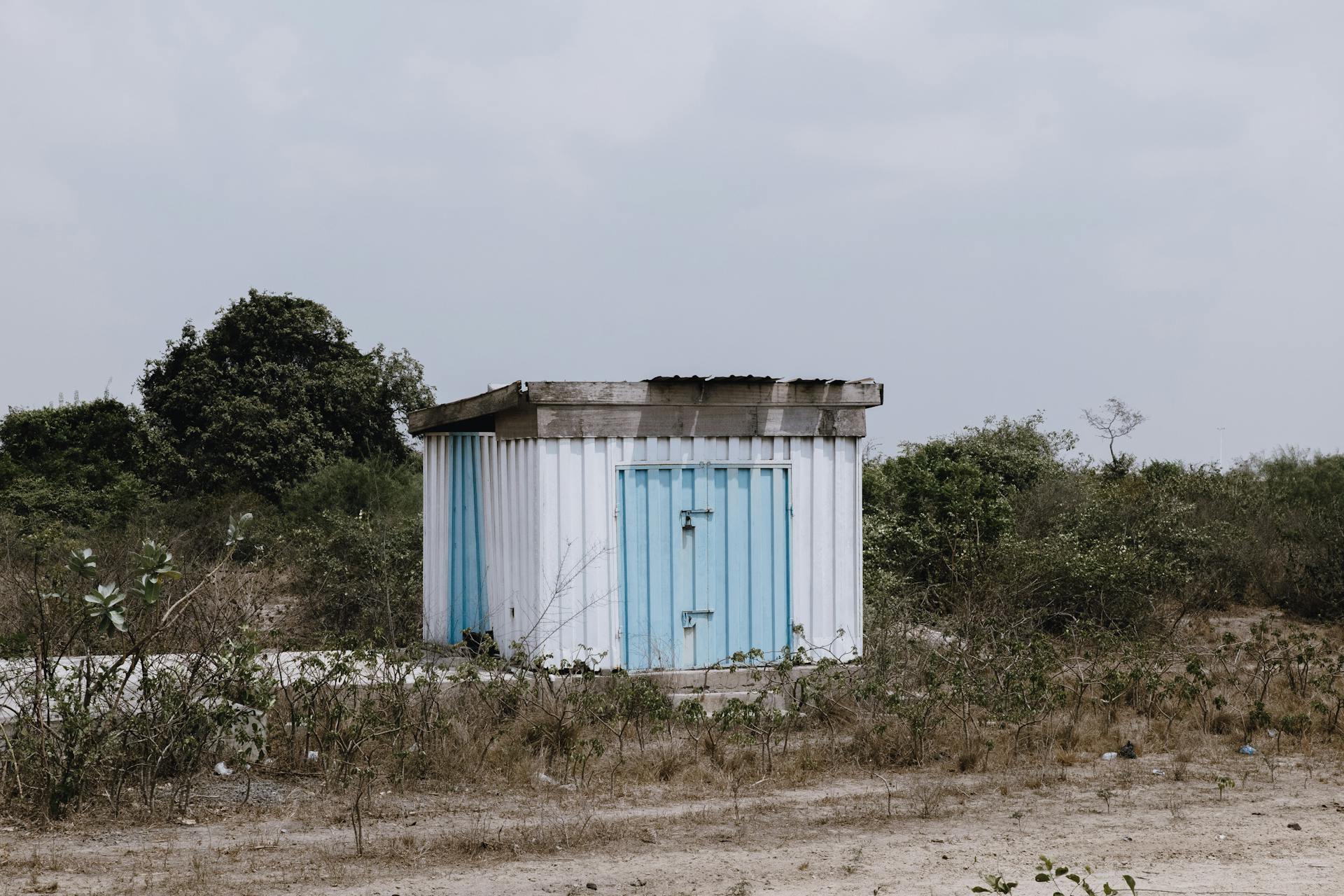 A small abandoned blue shed stands alone in a dry, rural landscape under a cloudy sky.