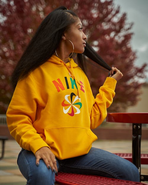 Selective Focus Photo of Woman in Yellow Hoodie and Blue Jeans Sitting on Red Bench While Looking Away