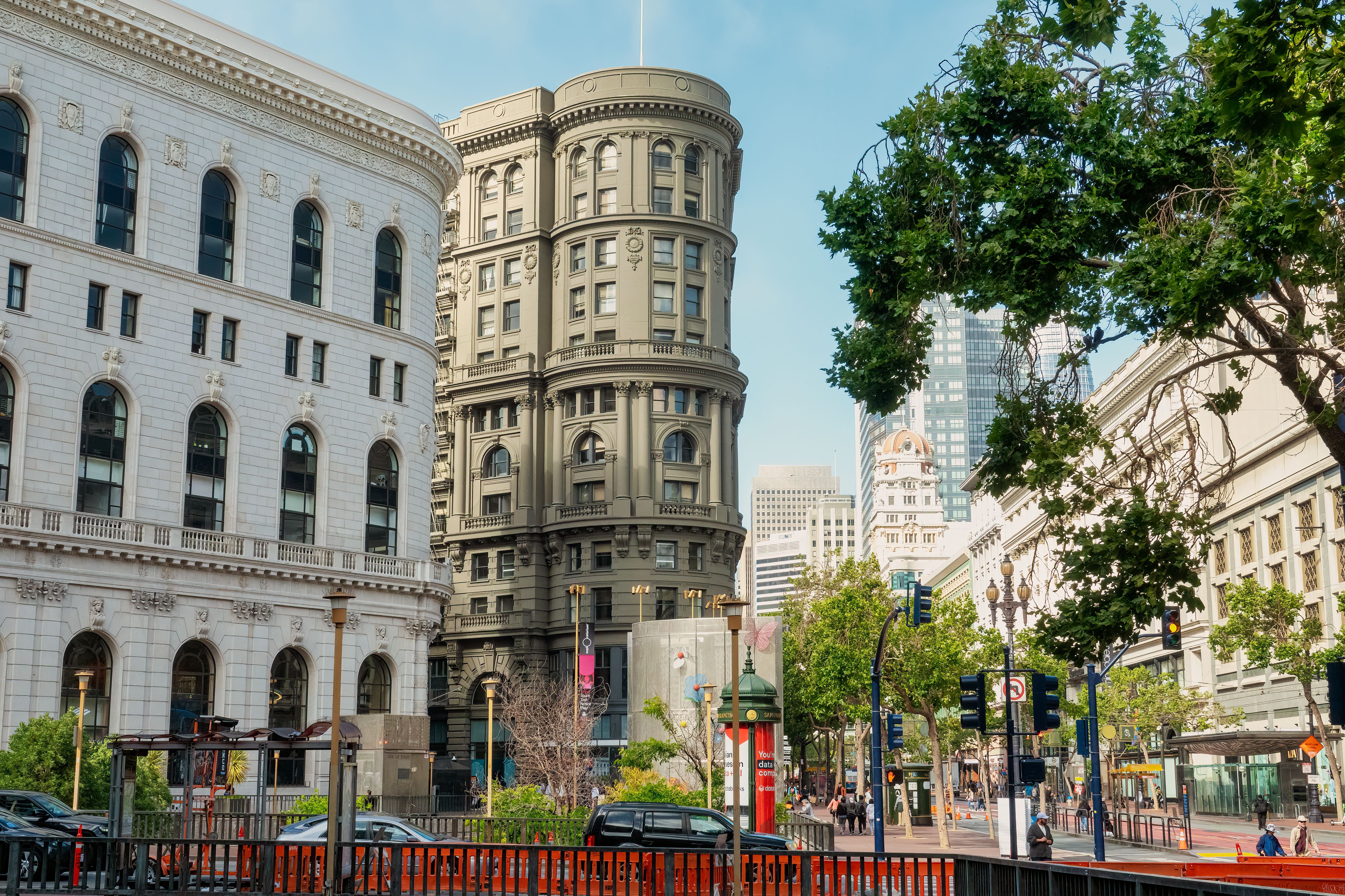 A curved stone-faced corner building between two classical buildings on a wide tree-lined street, with taller towers behind