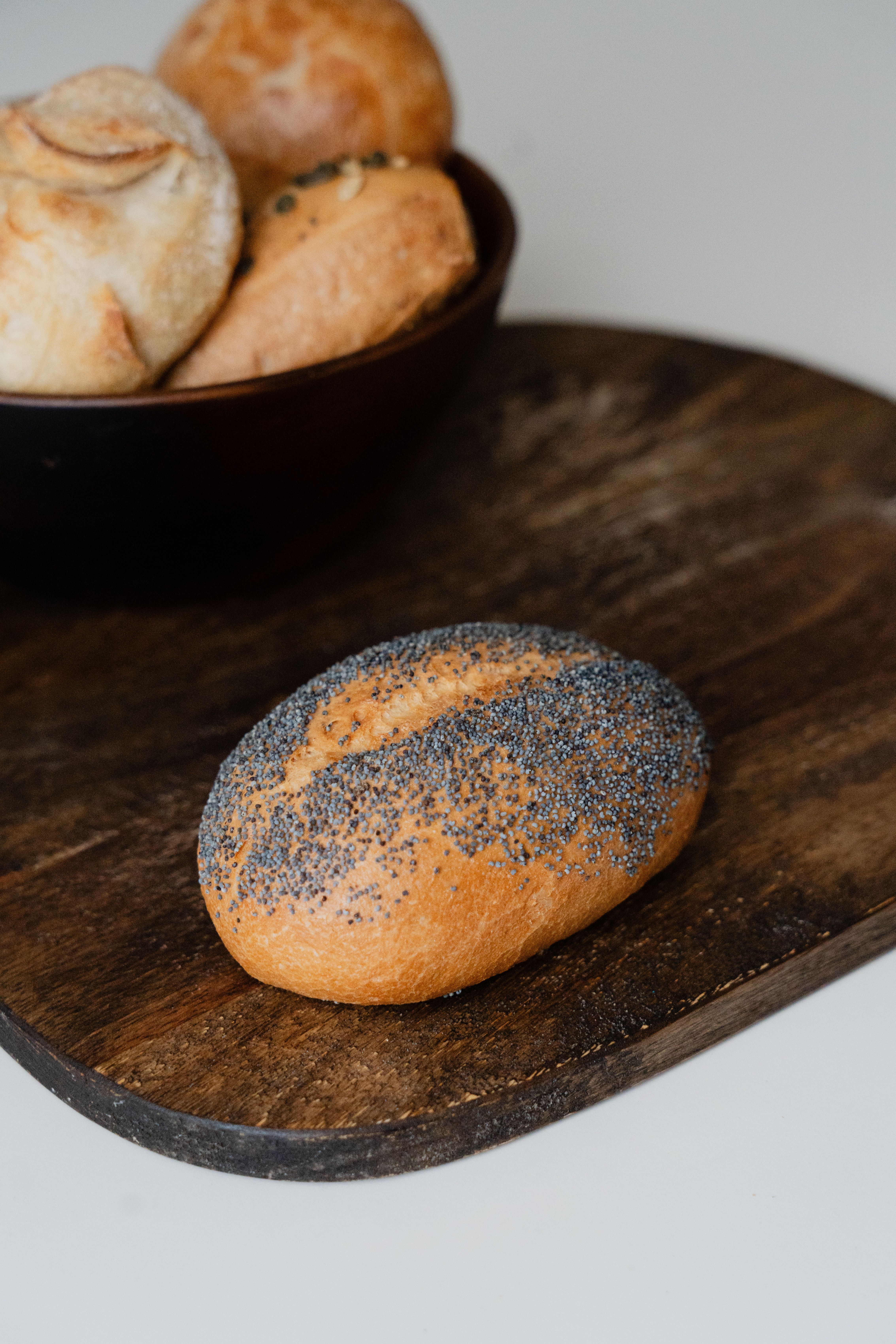 Close-up of a poppy seed bread roll on a wooden board next to a bowl of assorted breads.