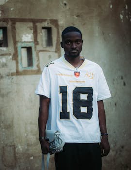 A man wearing a sports jersey stands against a textured wall, holding a bag.
