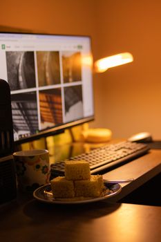Warm-lit desk setup with a monitor, cake, and floral mug in a cozy workspace.
