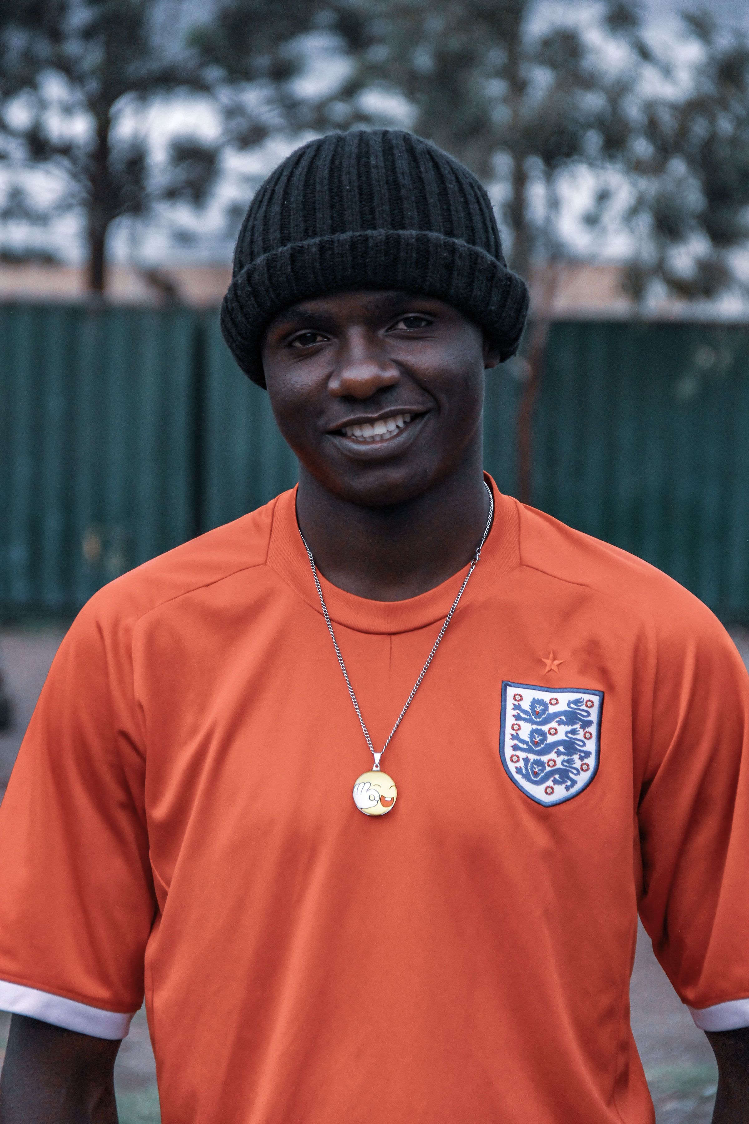 A young man smiles in a sports jersey with a gold necklace, captured outdoors.