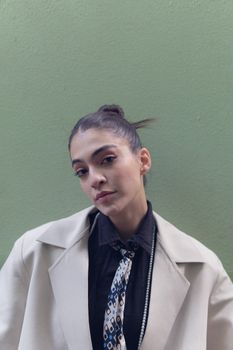 Stylish woman in trench coat and tie posing against a green wall.