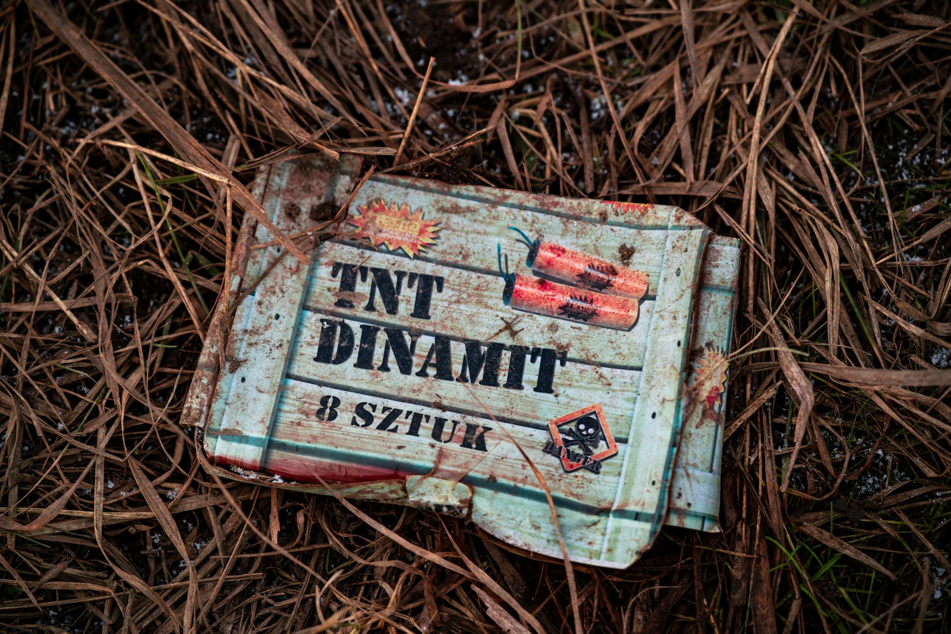 An old TNT box lies on dry grass, showcasing rustic and gritty textures outdoors.