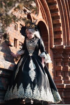 Woman in ornate black and gold gown standing by a historic red brick archway.