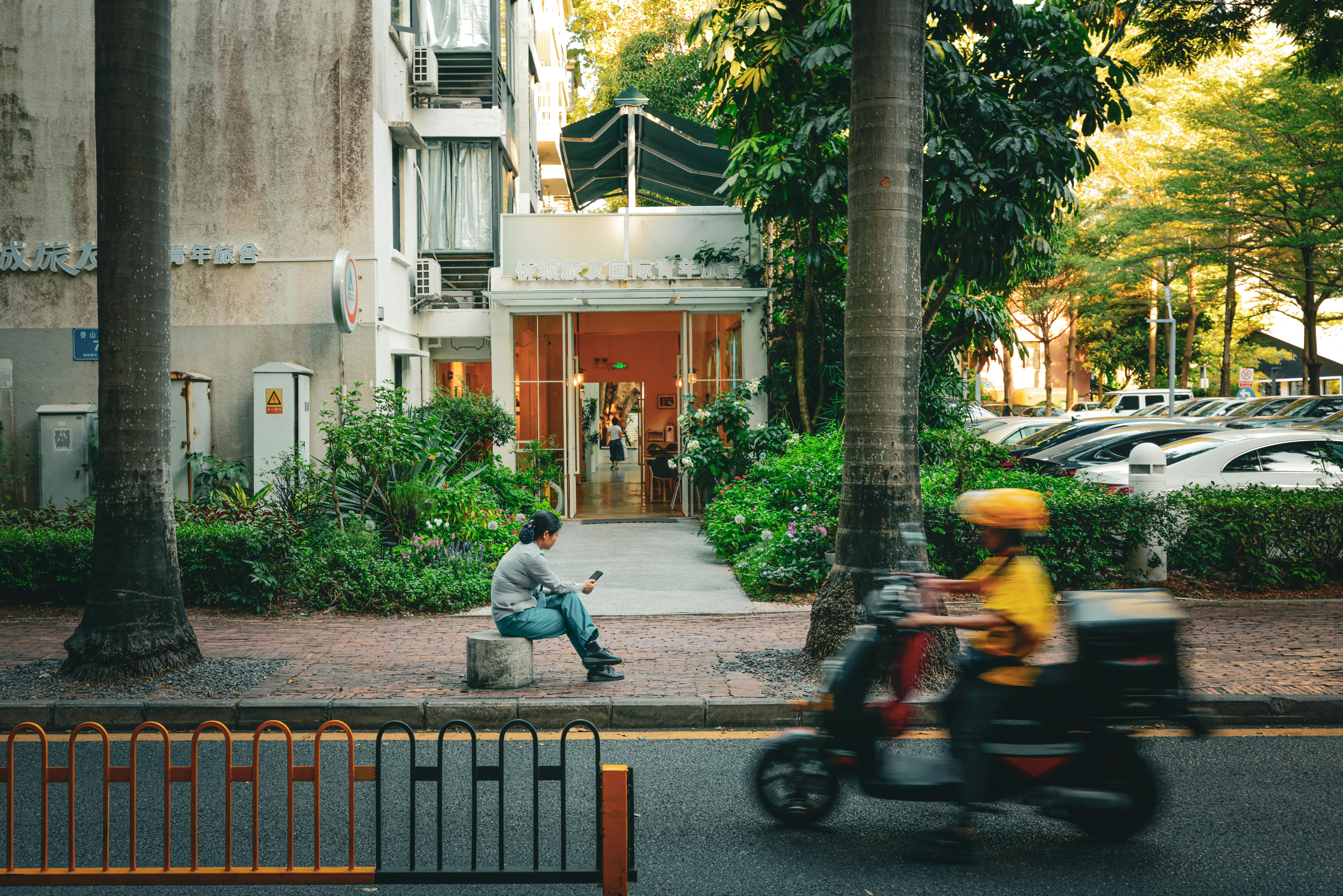 A man sits near a building while a scooter passes by on a sunny day in Shenzhen, China.