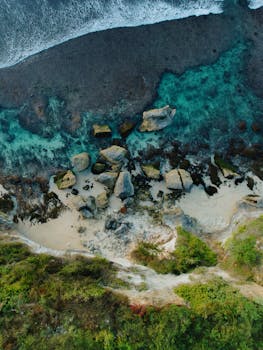 Stunning aerial view of Bali's coastline with clear waters, rocky shoreline, and lush greenery.