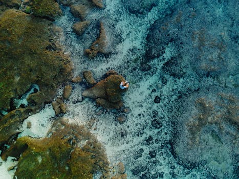 Aerial shot of a person on a rock surrounded by clear blue ocean in Bali, Indonesia.