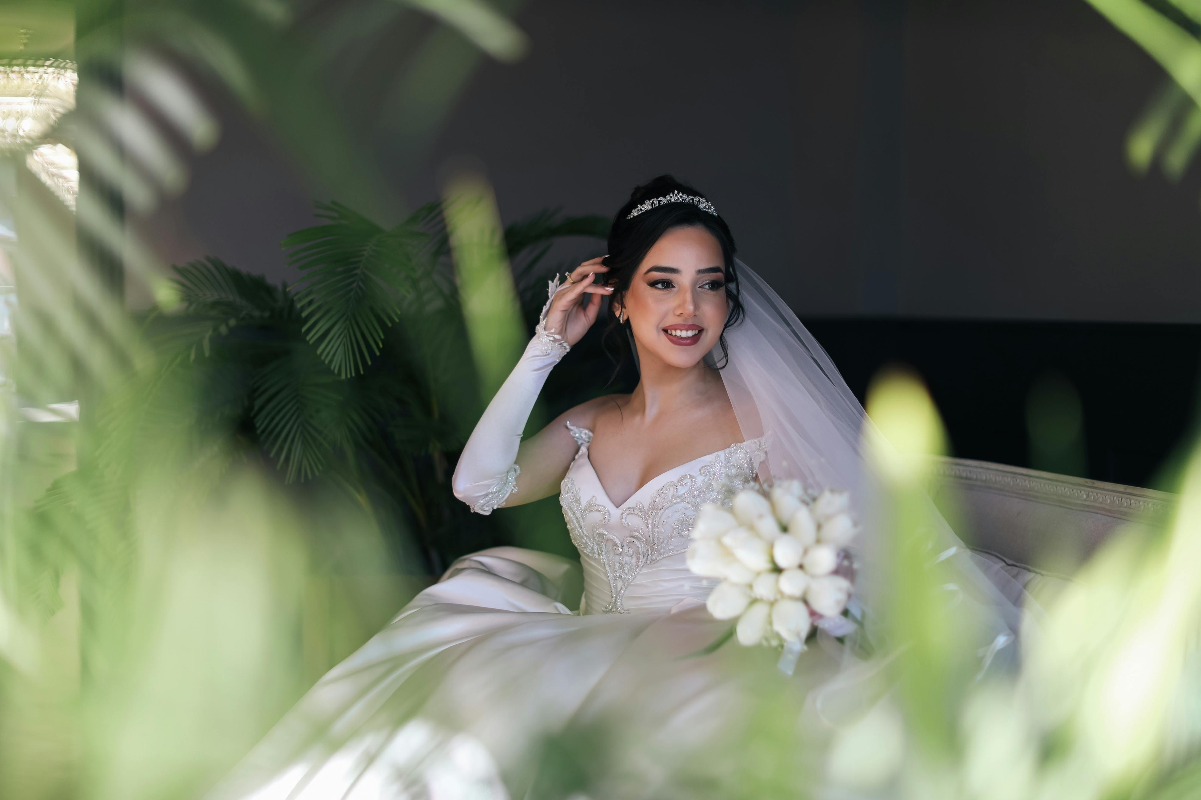 Bride in white gown and veil holding bouquet, smiling indoors.