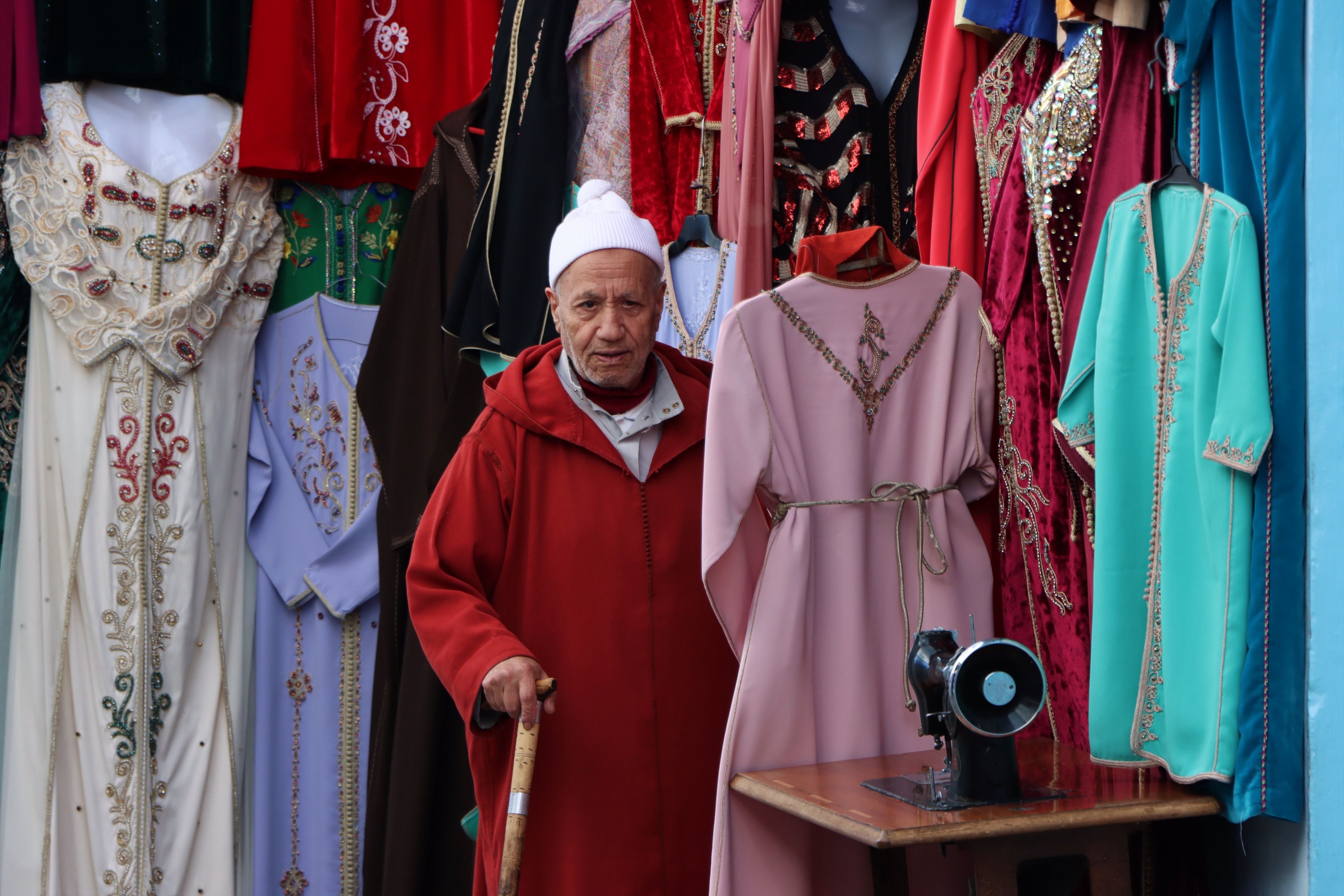 Elderly man in traditional attire at Moroccan clothing shop with colorful dresses.