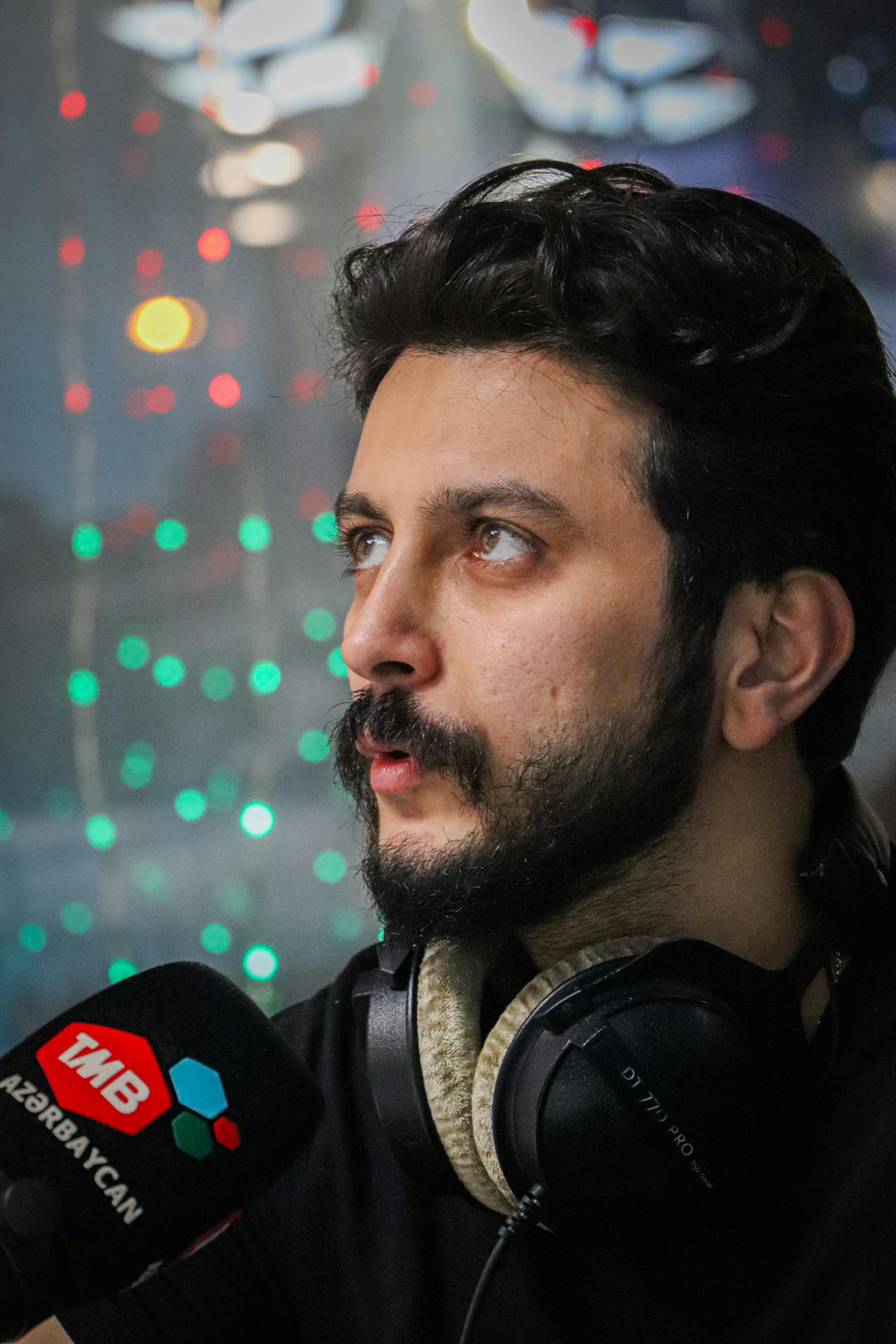 Man with beard speaking into a microphone wearing headphones with colorful lights in the background.
