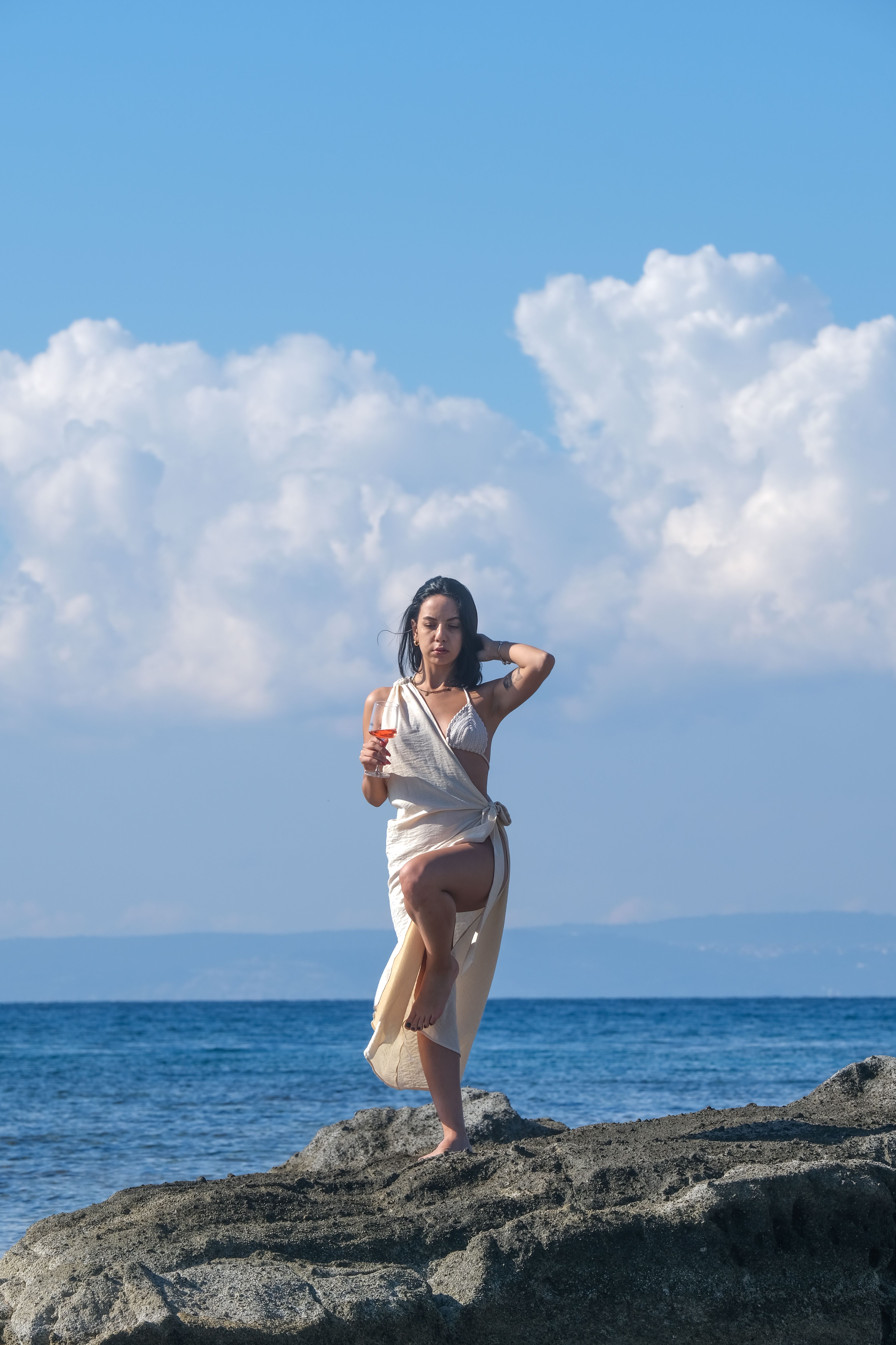 A stylish woman stands gracefully on rocks by the sea under a clear summer sky.
