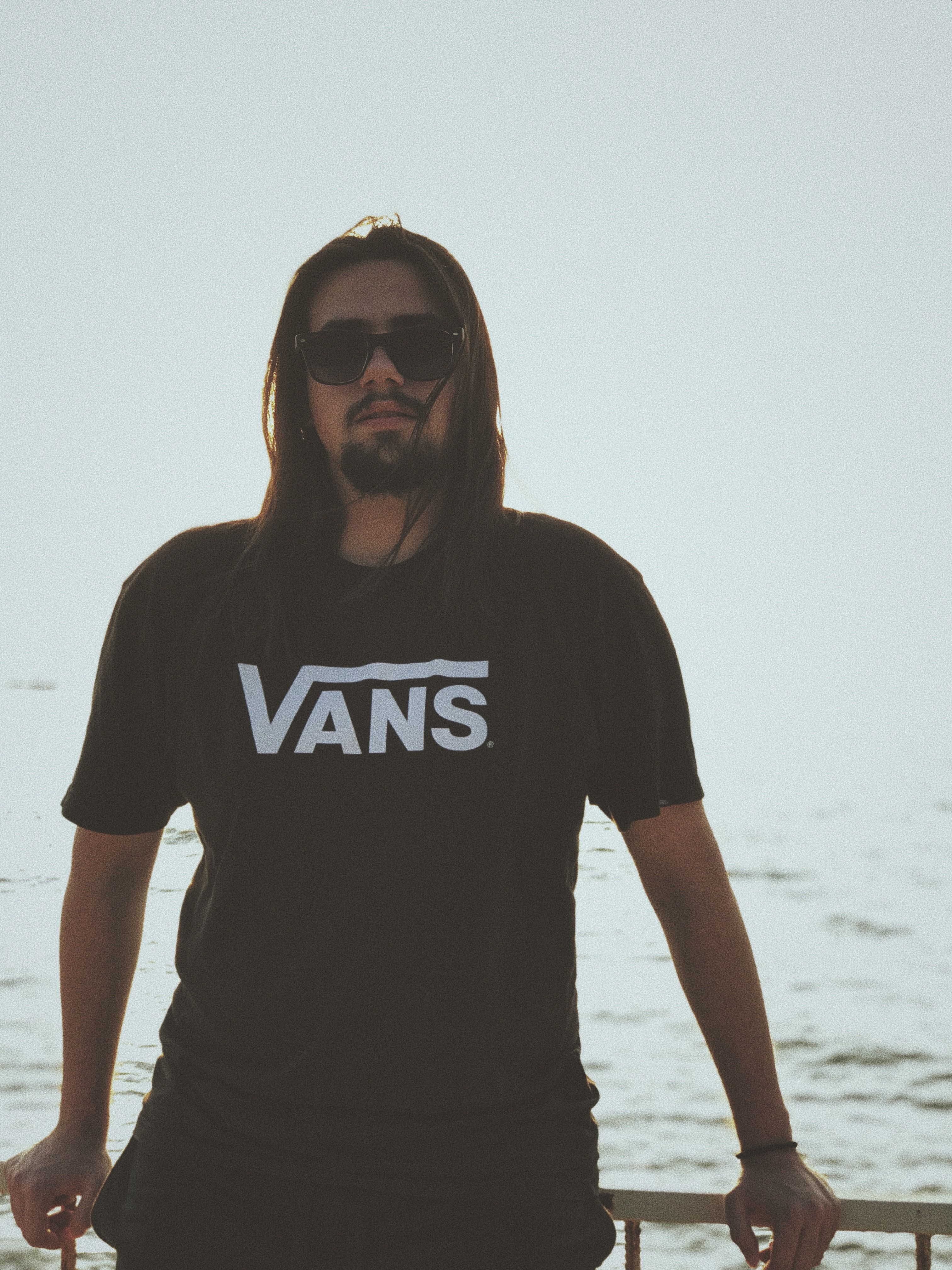 Man with sunglasses in Vans t-shirt standing outdoors by the sea.