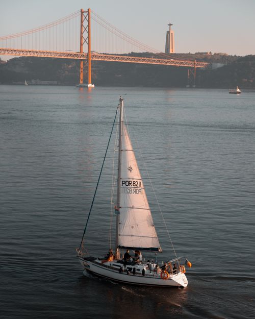 Free Sailboat sailing under the 25 de Abril Bridge on the Tagus River in Lisbon, Portugal, during a calm day. Stock Photo